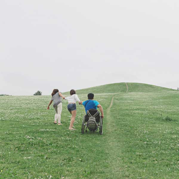As a group of friends engages in one of their favorite summer events, hiking a grassy hill, one of them is in a wheelchair and being pulled.